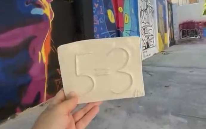 A 5=3 plaque held up in front of a colourful street-art mural in Cyprus