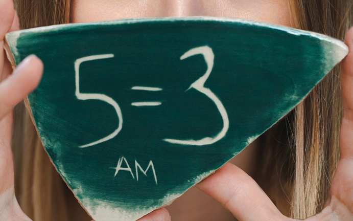 A close-up of the hand-painted 5=3 formula on a ceramic plaque, held up in front of Anna Maslennikov's face.
