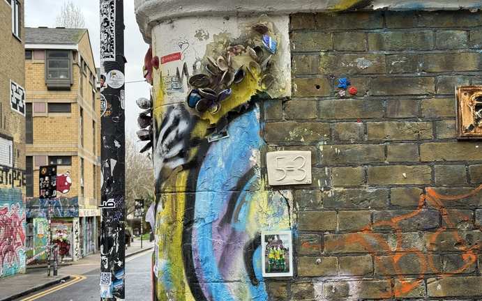 A ceramic 5=3 plaque mounted on a brick wall covered in colourful London street art