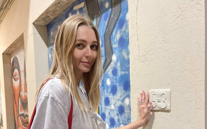 Anna Maslennikov beside a ceramic 5=3 plaque on a street-art wall in Tel Aviv.