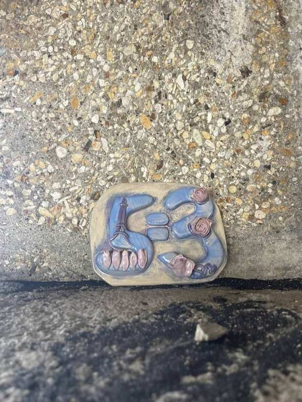 A small ceramic 5=3 plaque resting on gravel among ivy