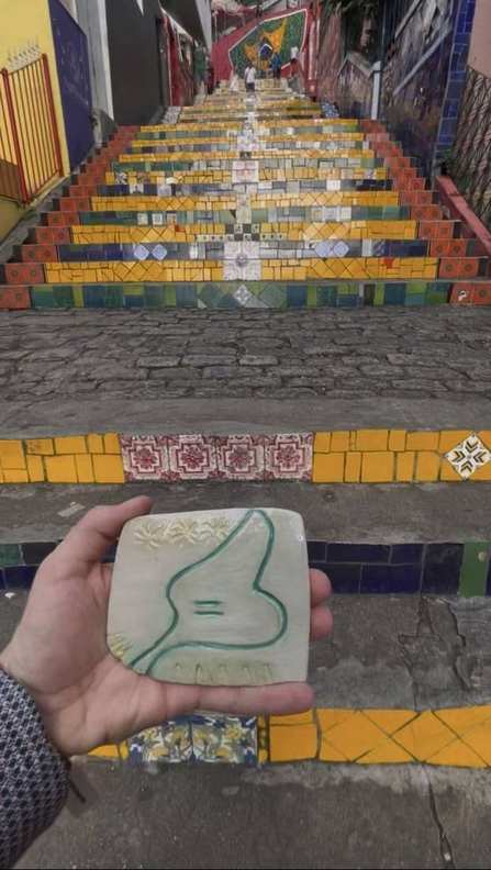 A hand-painted 5=3 ceramic tile held against a colourful tiled staircase
